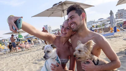 Coppia scatta selfie sulla spiaggia con due cani sotto ombrelloni