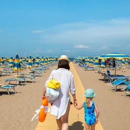 Donna e bambina che camminano su una spiaggia con ombrelloni blu e gialli