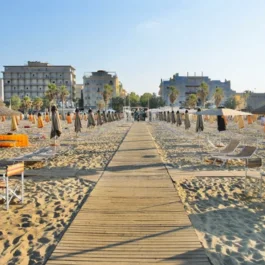Strand mit geschlossenen Sonnenschirmen, Liegestühlen und Holzsteg zur Stadt