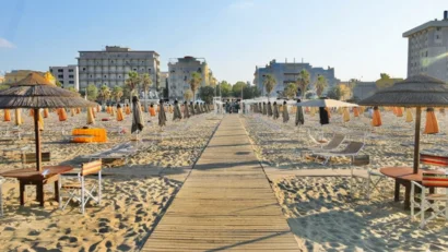 Strand mit geschlossenen Sonnenschirmen, Liegestühlen und Holzsteg zur Stadt
